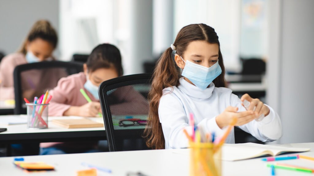 Small schoolgirl applying antibacterial sanitizer from small bottle, spraying it on palms, wearing disposable protective medical mask, sitting in classroom during lesson